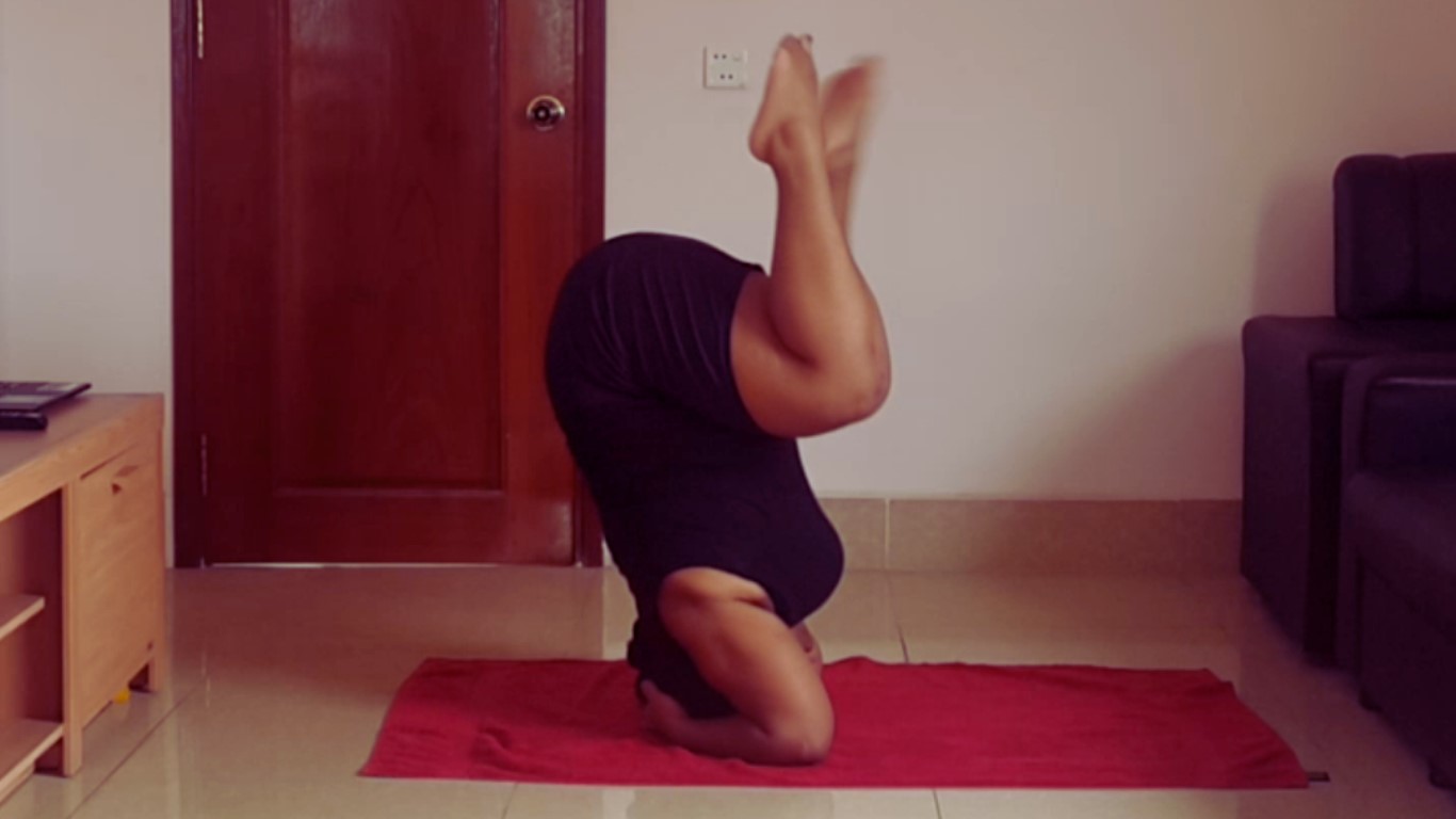 Agneatha practising a headstand yoga pose indoors on a red towel, with their legs bent and lifted in the air.
